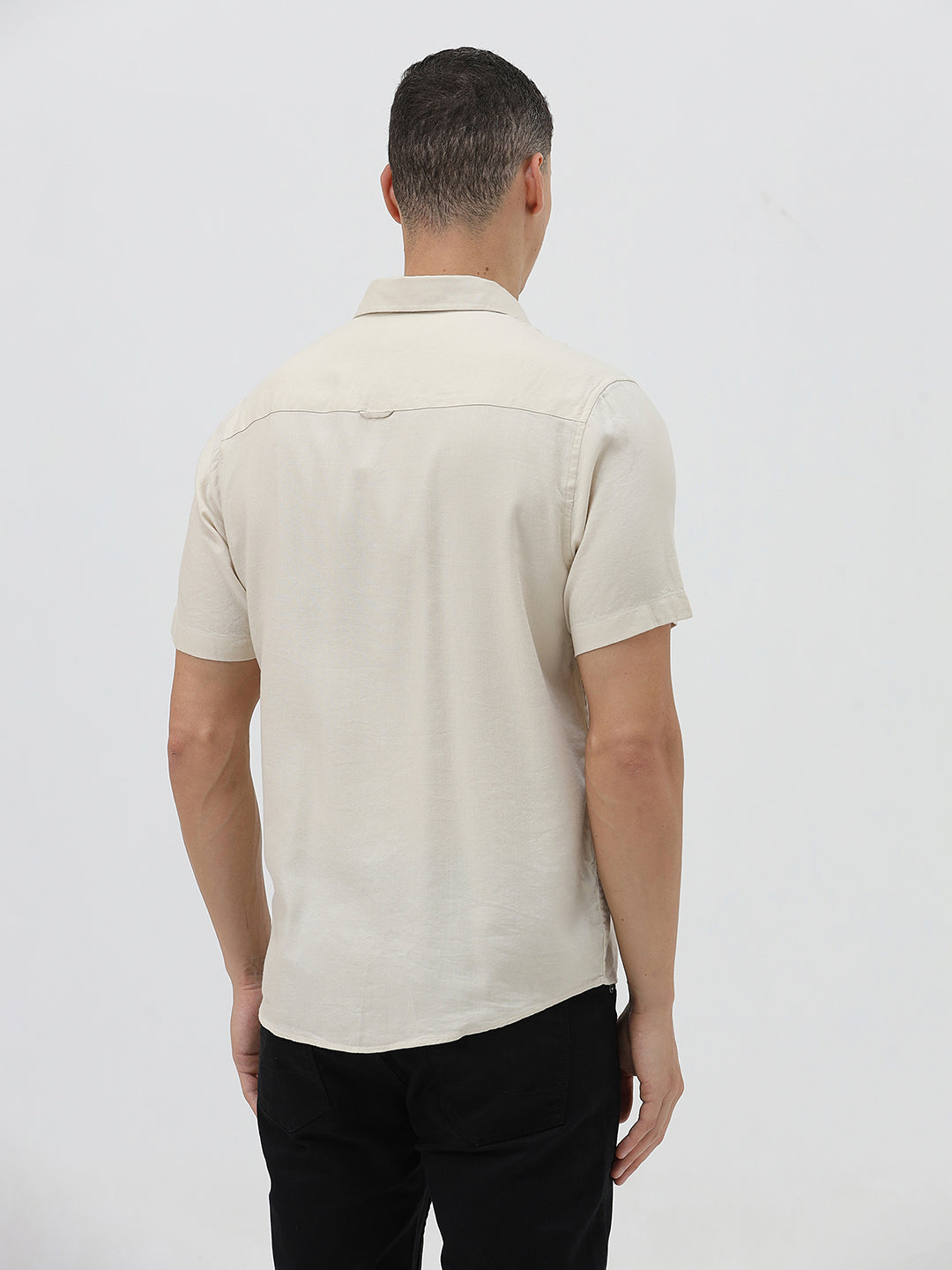 A man wearing a short-sleeve linen button-up shirt with a color-blocked design in beige and cream, paired with black pants, against a plain white studio background.