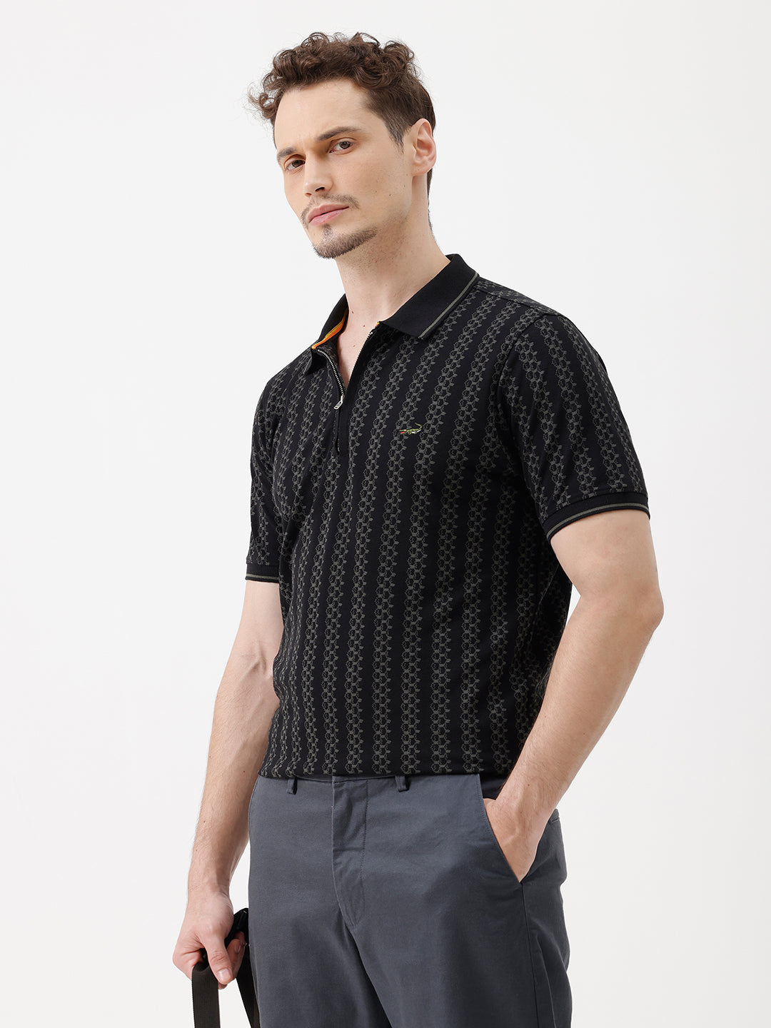 A man wearing a black short-sleeve polo shirt with a subtle geometric pattern, paired with dark gray trousers, standing against a plain white studio background.