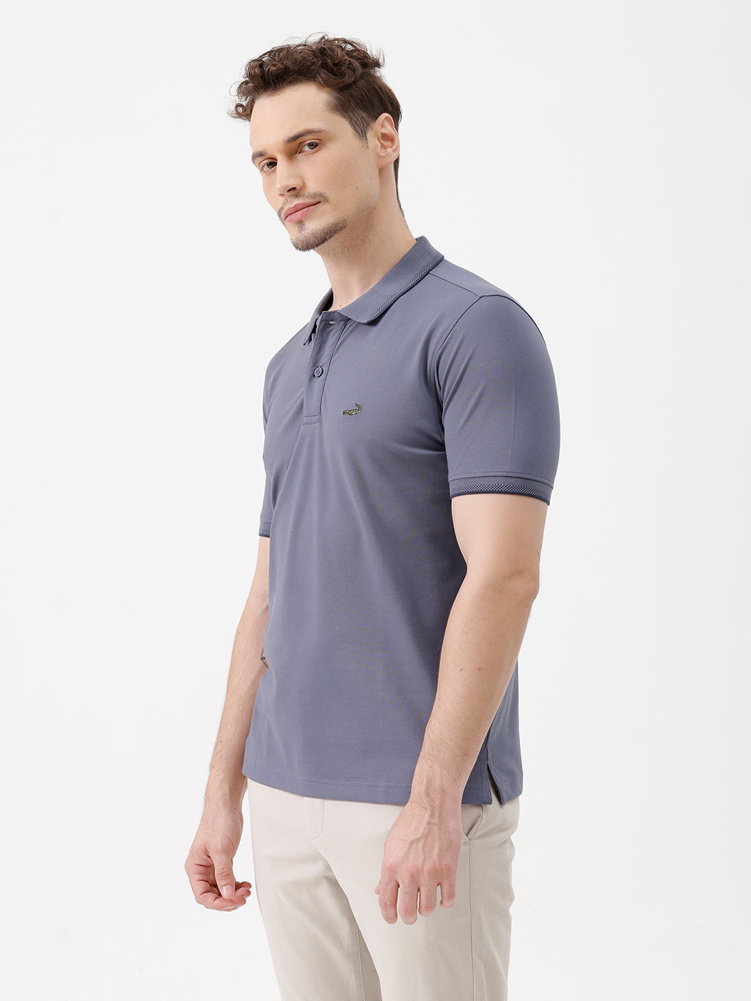 A man wearing a slate blue polo shirt and beige trousers, standing against a plain white background.