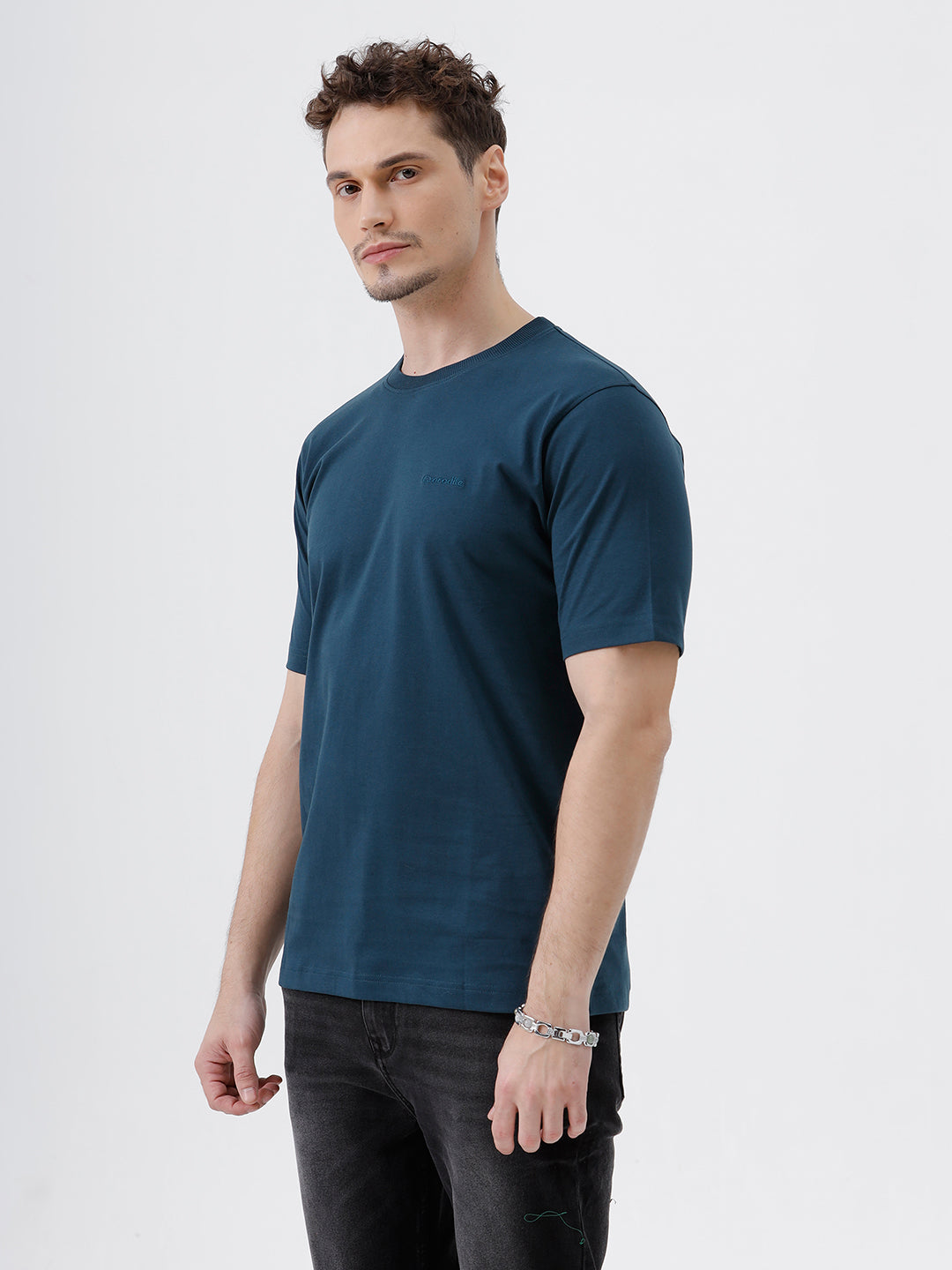 A man wearing a dark navy blue crew-neck t-shirt and black jeans, standing against a light gray studio background.