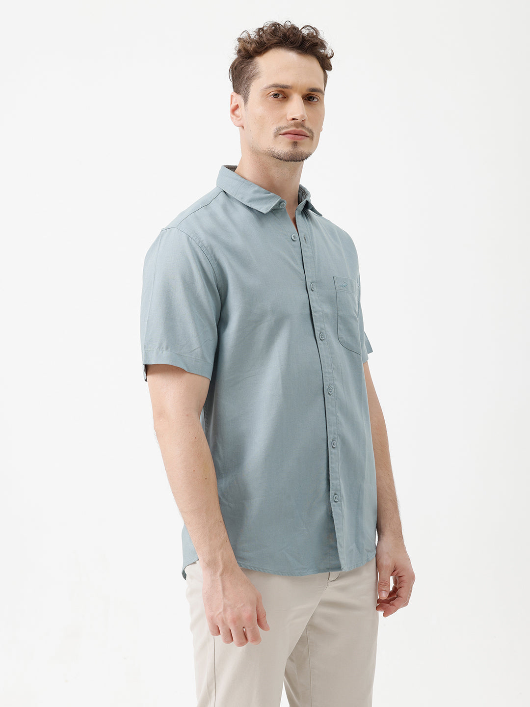 A man wearing a light blue short-sleeve button-up shirt and beige trousers, standing against a plain white studio background.