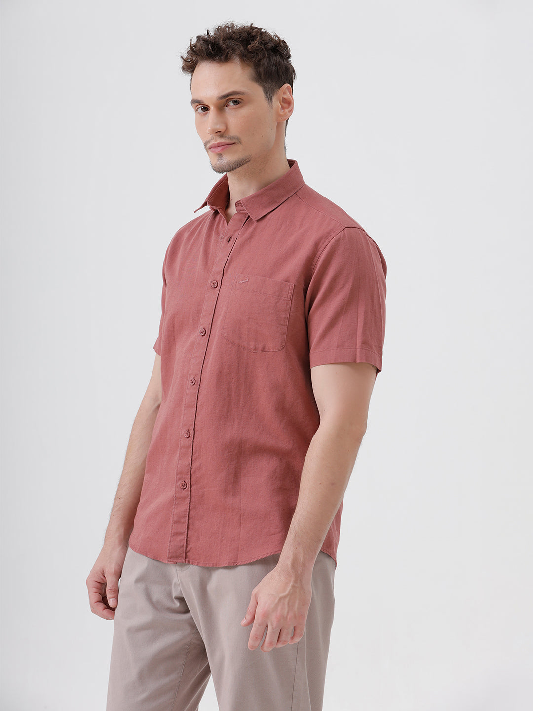A man wearing a short-sleeve button-up shirt in a dusty rose color, paired with beige trousers, standing against a light gray studio background.