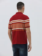 A man wearing a red short-sleeve polo shirt with horizontal cream and rust-orange stripes across the chest and sleeves, paired with dark blue jeans and black sunglasses, against a light gray studio background.