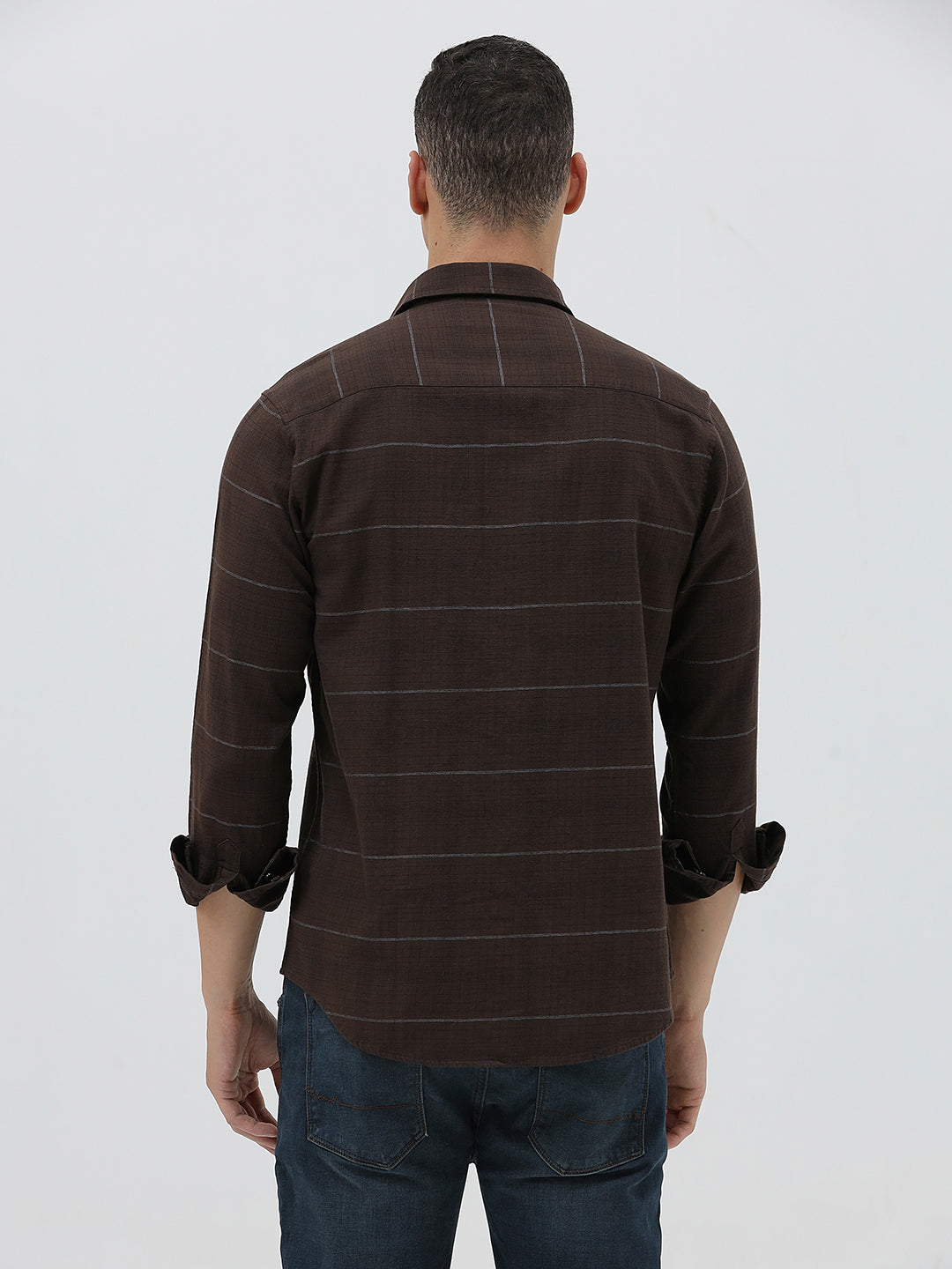 A man wearing a dark brown long-sleeve button-up shirt with thin horizontal stripes, paired with dark blue jeans, standing against a light gray studio background.