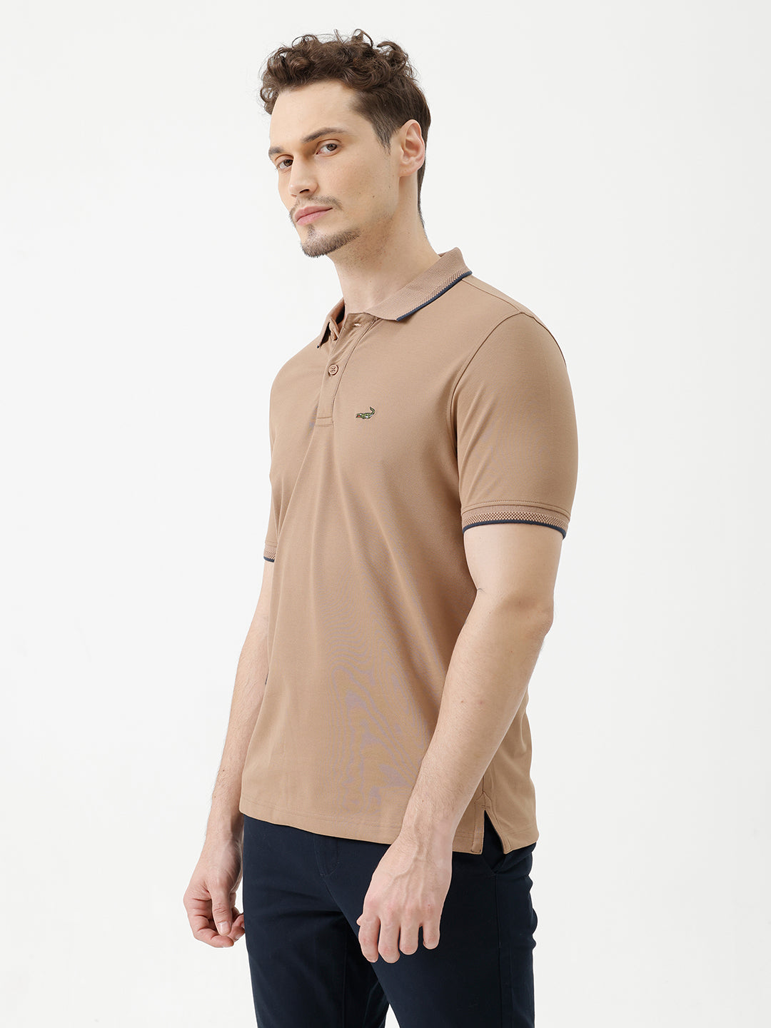 A man wearing a tan short-sleeve polo shirt with dark blue piping on the collar and sleeve cuffs, paired with dark navy pants, against a plain light gray background.
