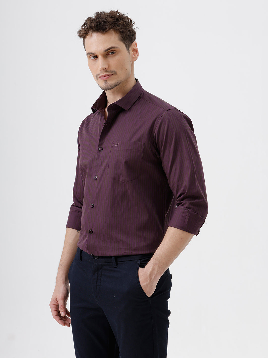 A man wearing a dark purple long-sleeve button-down shirt with subtle vertical stripes and dark navy trousers, standing against a light gray background.