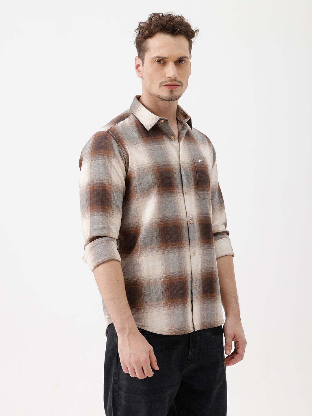 A man wearing a long-sleeve button-up plaid shirt in brown, beige, and gray tones, paired with dark jeans, standing against a plain white studio background.