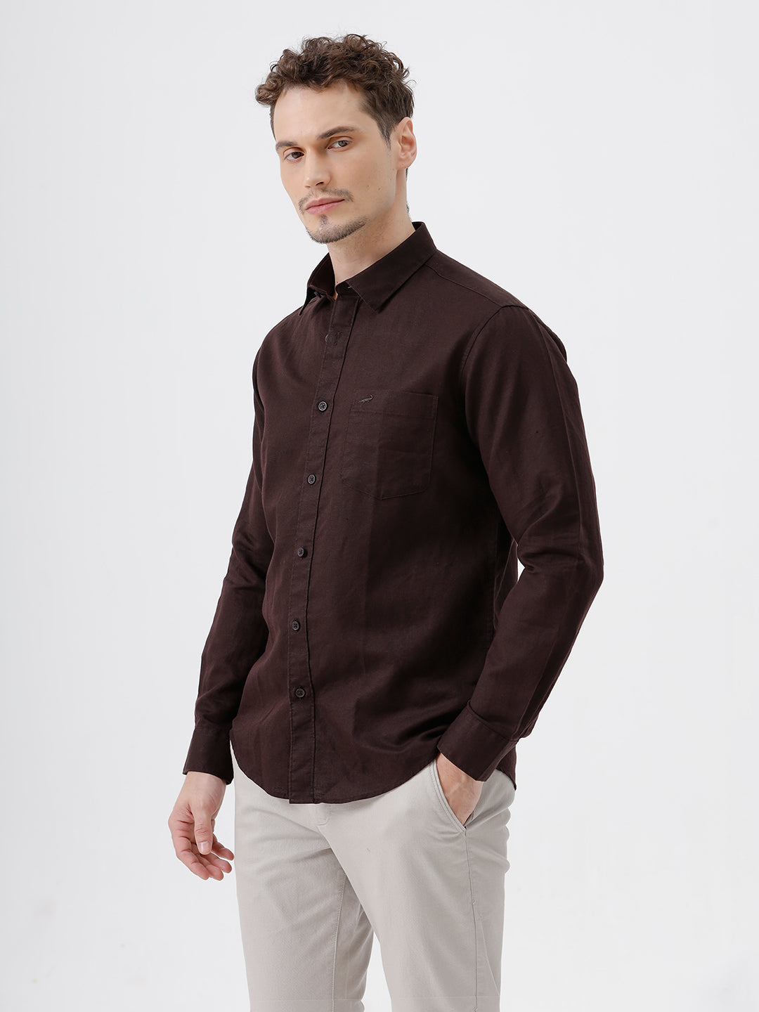A man wearing a dark brown long-sleeve button-down shirt and light beige trousers, standing against a plain light gray studio background.