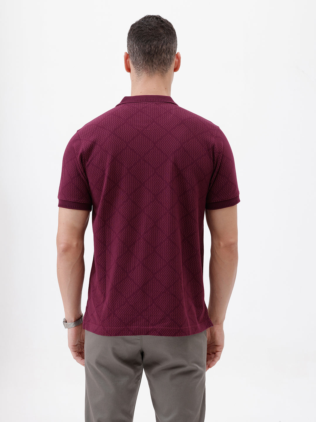 A man wearing a short-sleeve polo shirt in deep burgundy with a subtle textured geometric pattern, styled with gray trousers, sunglasses, and a watch against a plain white background.