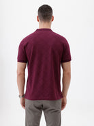 A man wearing a short-sleeve polo shirt in deep burgundy with a subtle textured geometric pattern, styled with gray trousers, sunglasses, and a watch against a plain white background.