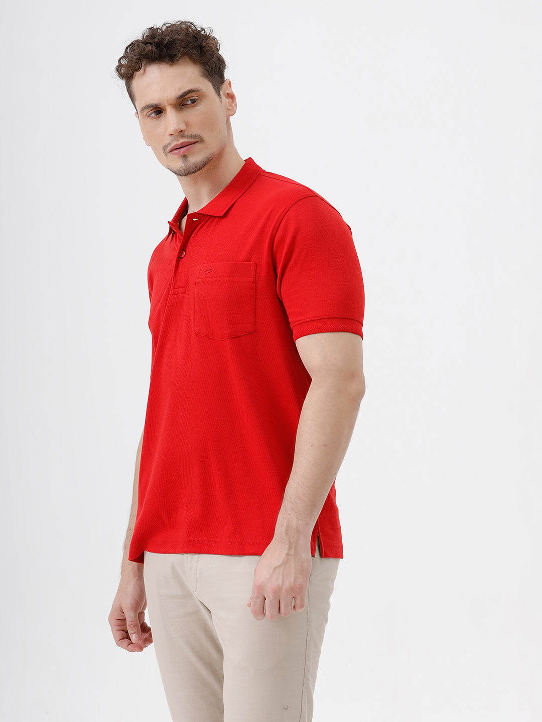 A man wearing a bright red short-sleeve polo shirt with a chest pocket, paired with beige trousers, standing against a plain white studio background.