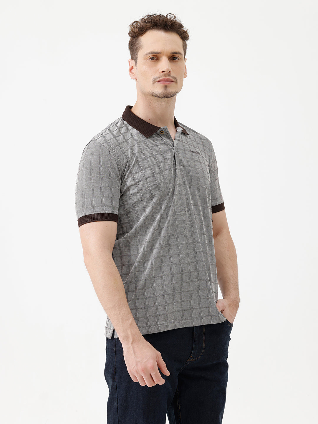 A man wearing a gray short-sleeve polo shirt with a subtle grid pattern and dark brown collar and sleeve trim, paired with dark blue jeans, against a plain white studio background.
