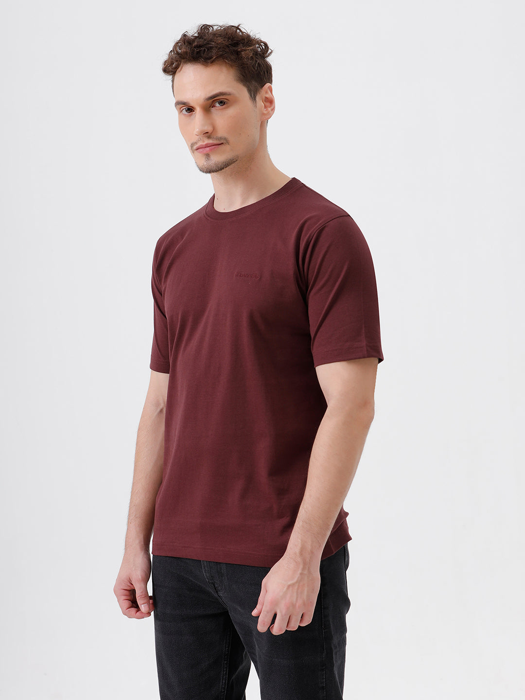 A man wearing a short-sleeve crewneck t-shirt in maroon, paired with dark jeans, standing against a plain light gray studio background.