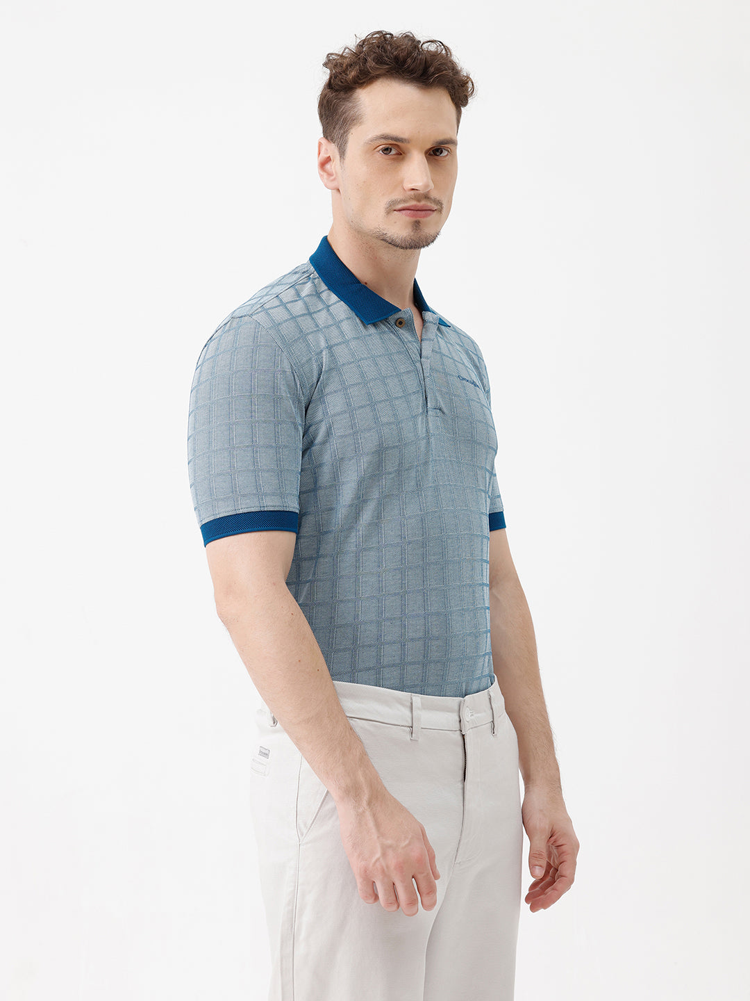 A man wearing a light blue and white checkered polo shirt with a dark blue collar and sleeve cuffs, paired with white trousers, standing against a plain white background.