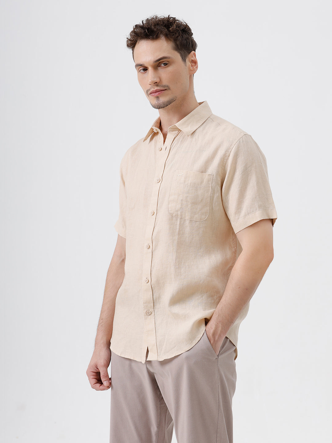 A man wearing a light beige short-sleeve linen button-up shirt and taupe trousers, standing against a plain white studio background.