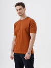 A man wearing a rust-orange crew-neck t-shirt and dark blue jeans, standing against a light gray studio background.