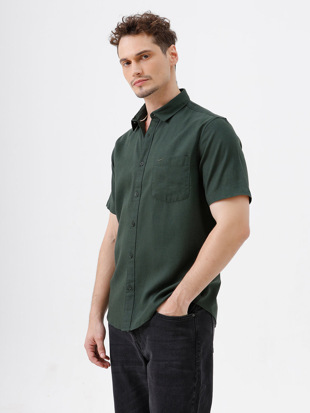 A man wearing a dark forest green short-sleeve button-up shirt with a chest pocket, paired with black jeans, standing against a plain white studio background.