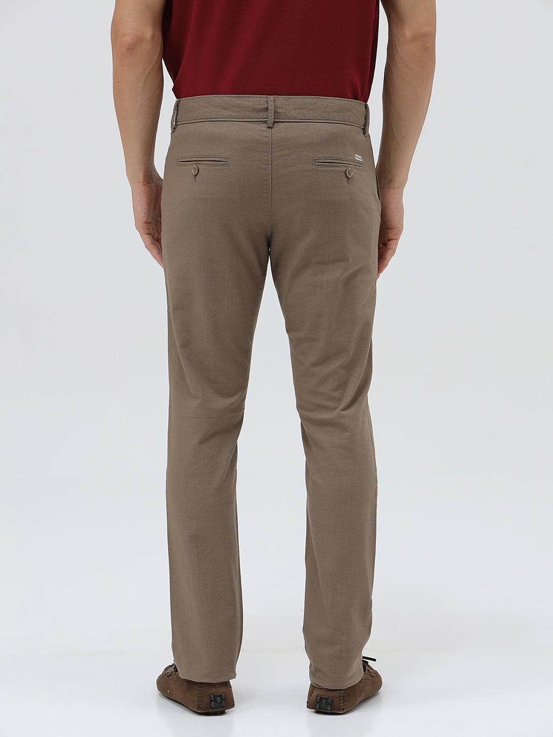 A person wearing slim-fit brown trousers, paired with a dark red shirt and brown suede moccasin shoes, shown from the waist down against a plain white studio background.