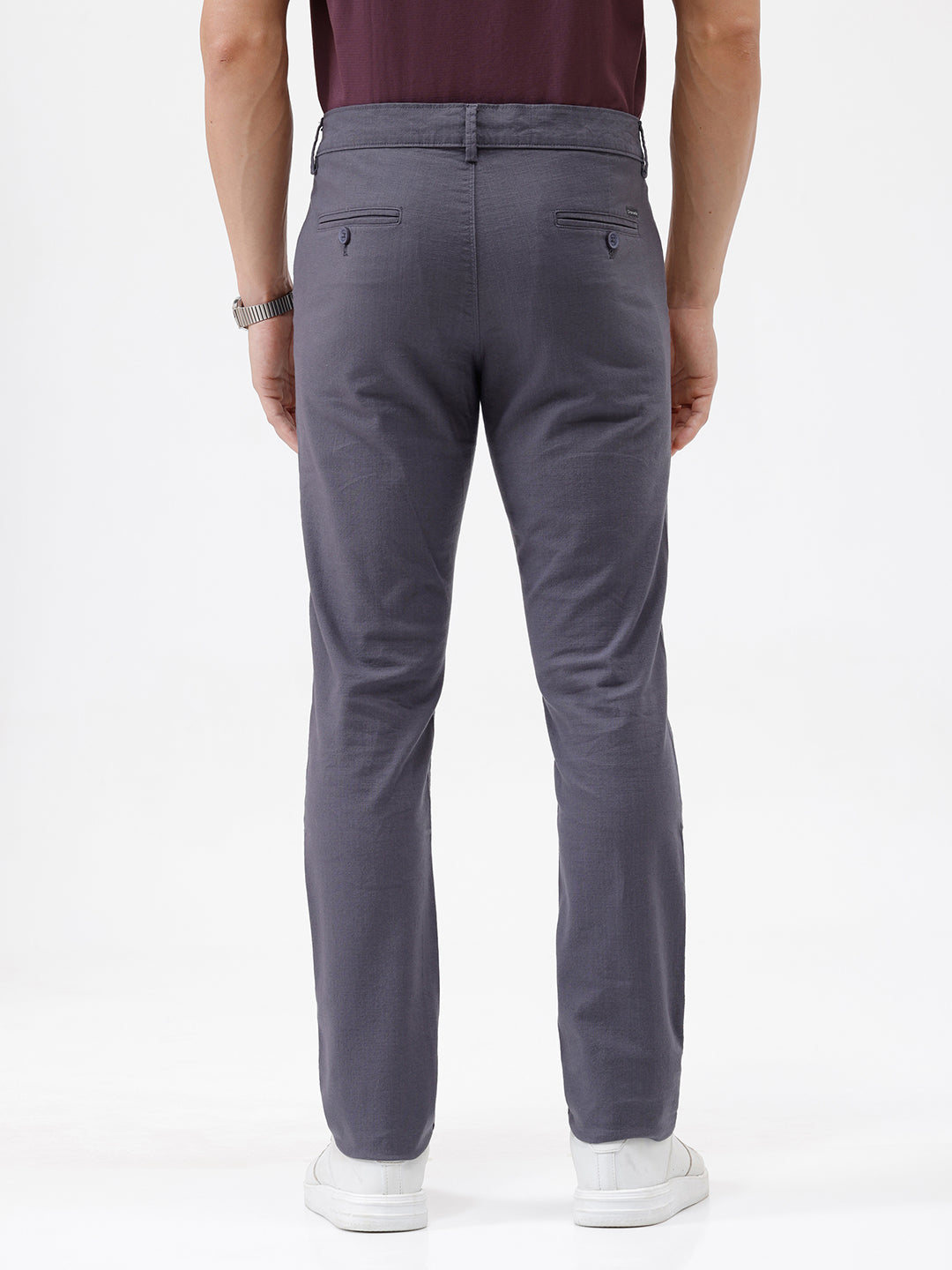 A person wearing slim-fit charcoal gray chino pants, paired with a dark purple shirt and white sneakers, standing against a plain white studio background.