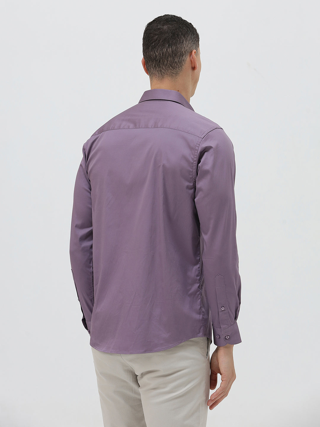 A man wearing a purple long-sleeve button-down shirt with rolled-up sleeves and light beige trousers, standing against a plain light gray background.
