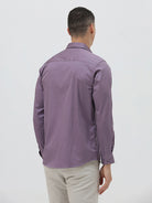 A man wearing a purple long-sleeve button-down shirt with rolled-up sleeves and light beige trousers, standing against a plain light gray background.