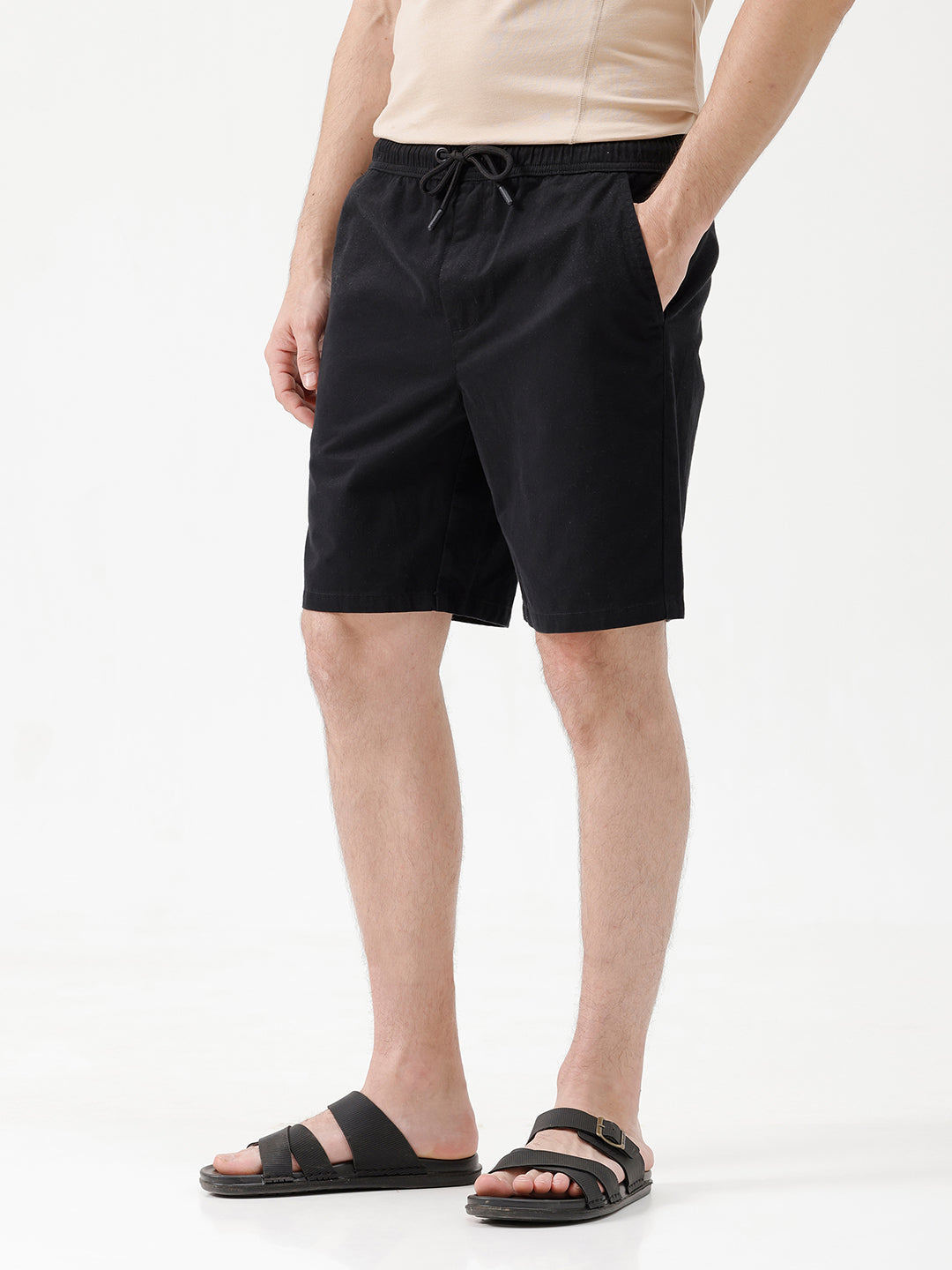 A man modeling a light beige short-sleeve t-shirt, black drawstring shorts, and black slide sandals against a plain white studio background.