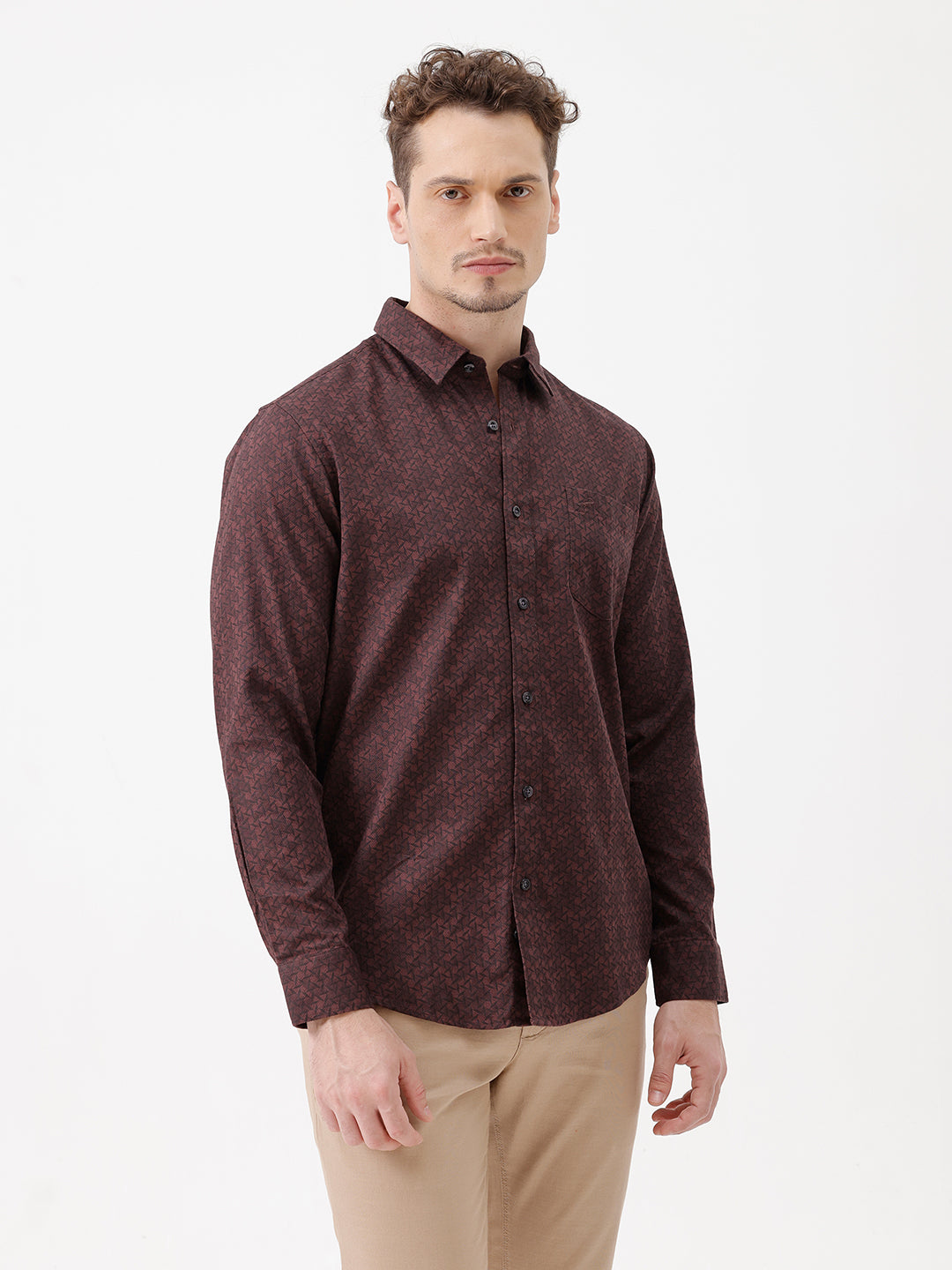A man wearing a dark burgundy long-sleeve button-up shirt with a subtle geometric pattern, paired with beige trousers, standing against a plain white studio background.
