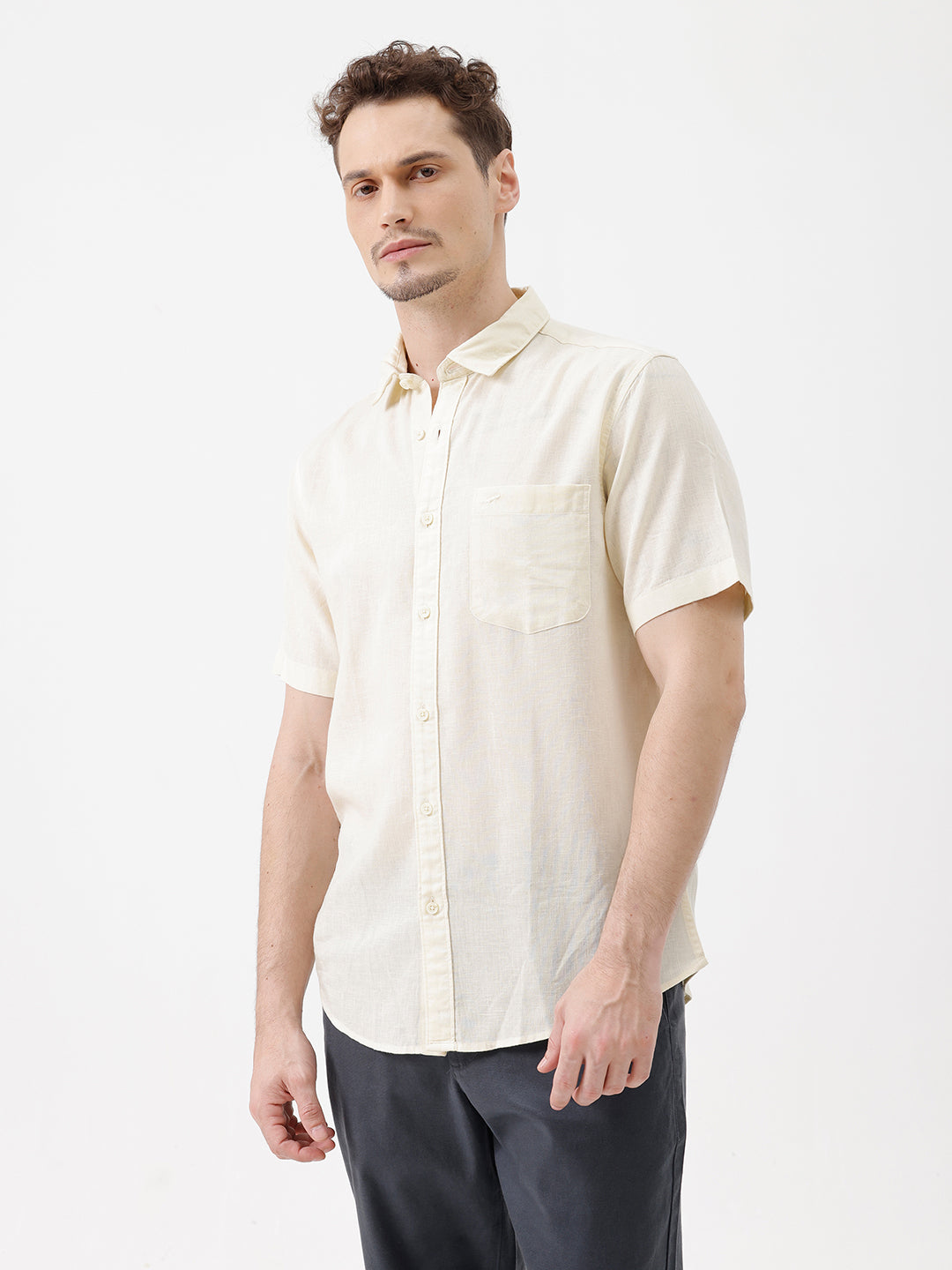 A man wearing a light cream-colored short-sleeve linen button-up shirt with a chest pocket, paired with dark trousers, against a plain white studio background.