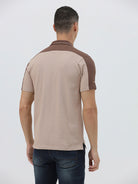 A man modeling a short-sleeve polo shirt in beige with brown shoulder panels and collar trim, paired with dark blue jeans, against a light gray studio background.
