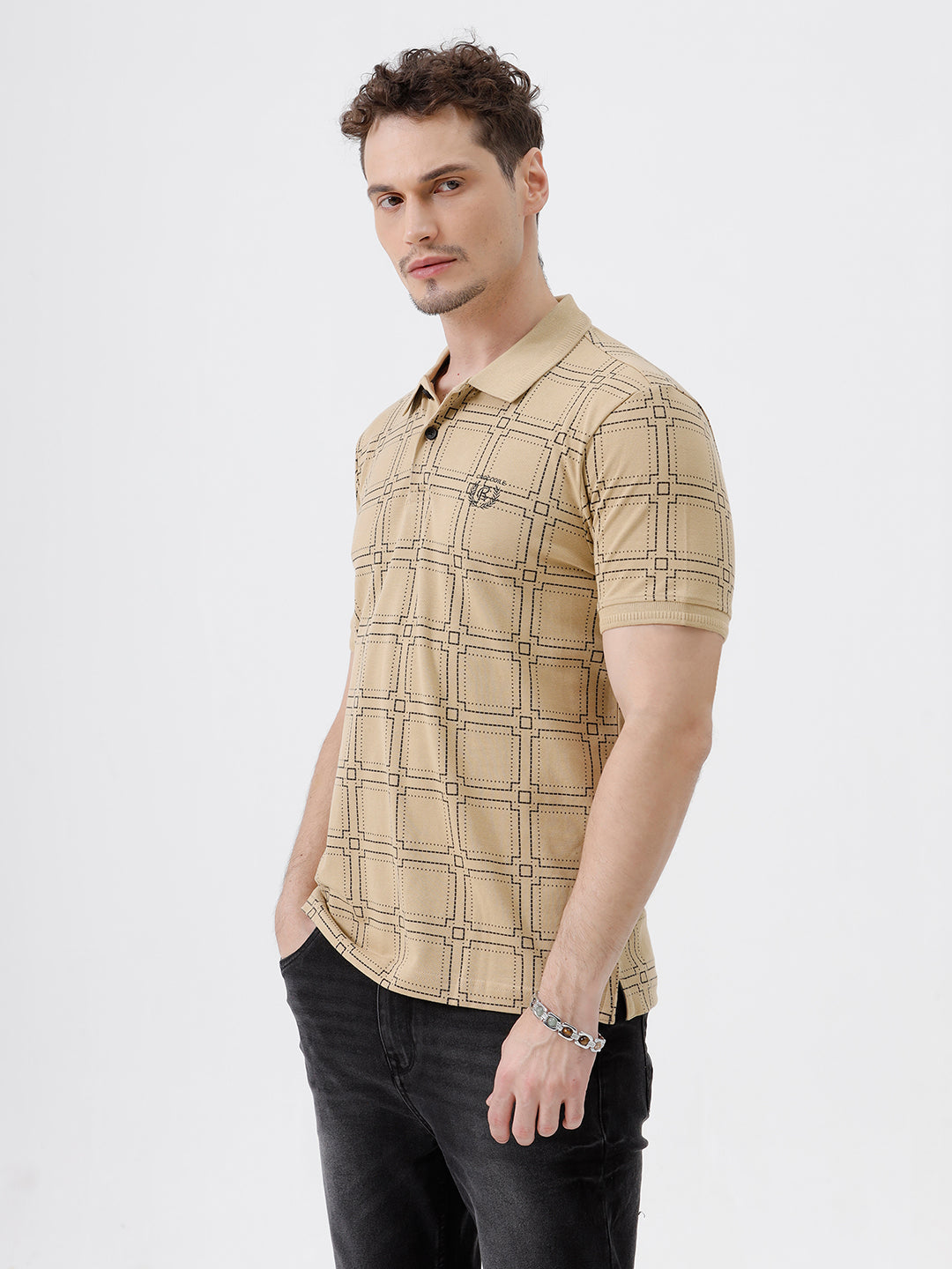 A man wearing a beige short-sleeve polo shirt with a brown grid pattern, paired with black jeans, standing against a plain white studio background.