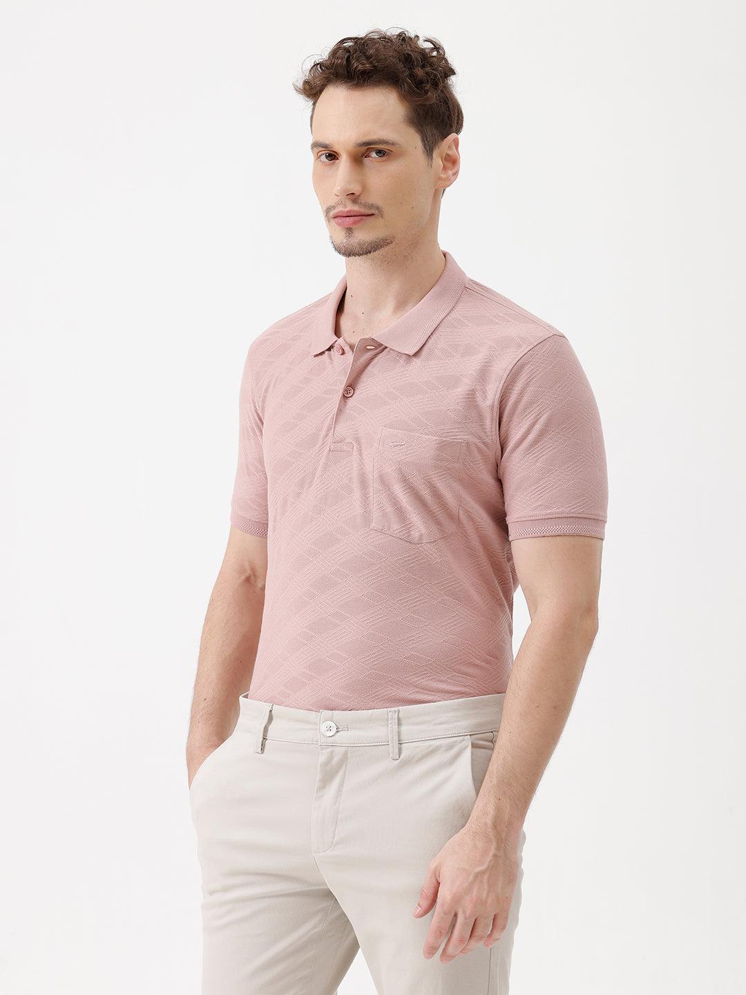 A man wearing a dusty pink short-sleeve polo shirt with a chest pocket, paired with light beige trousers, standing against a plain white studio background.