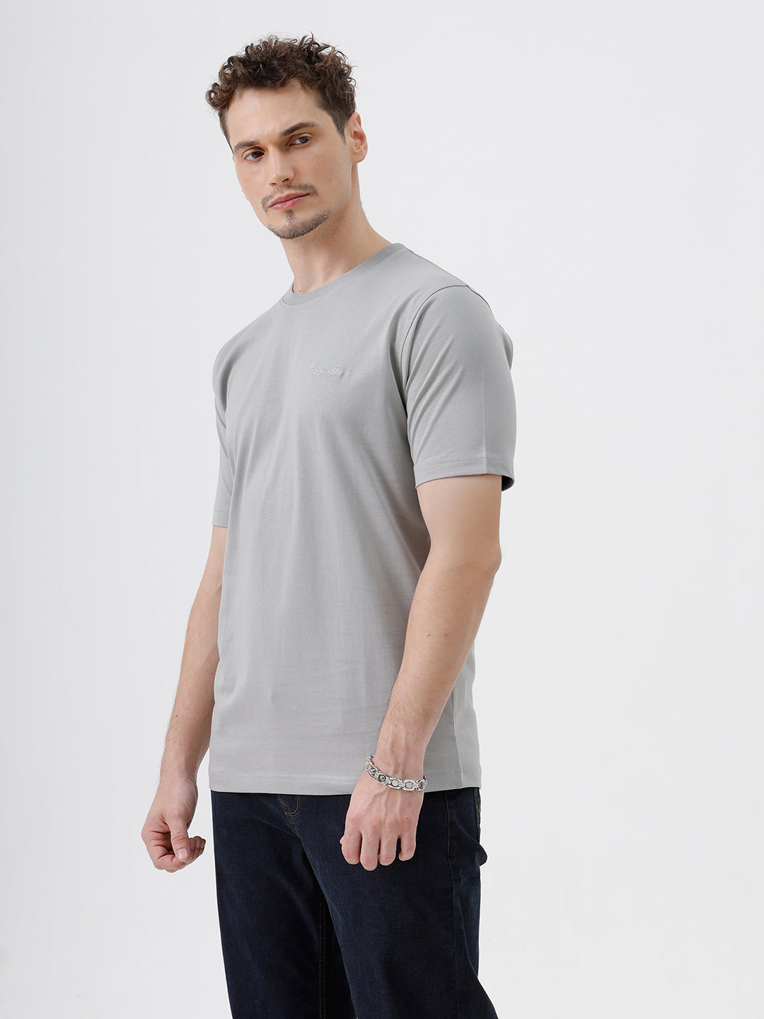 A man wearing a light gray short-sleeve crewneck t-shirt with a subtle tonal chest logo, paired with dark jeans, standing against a light gray studio background.