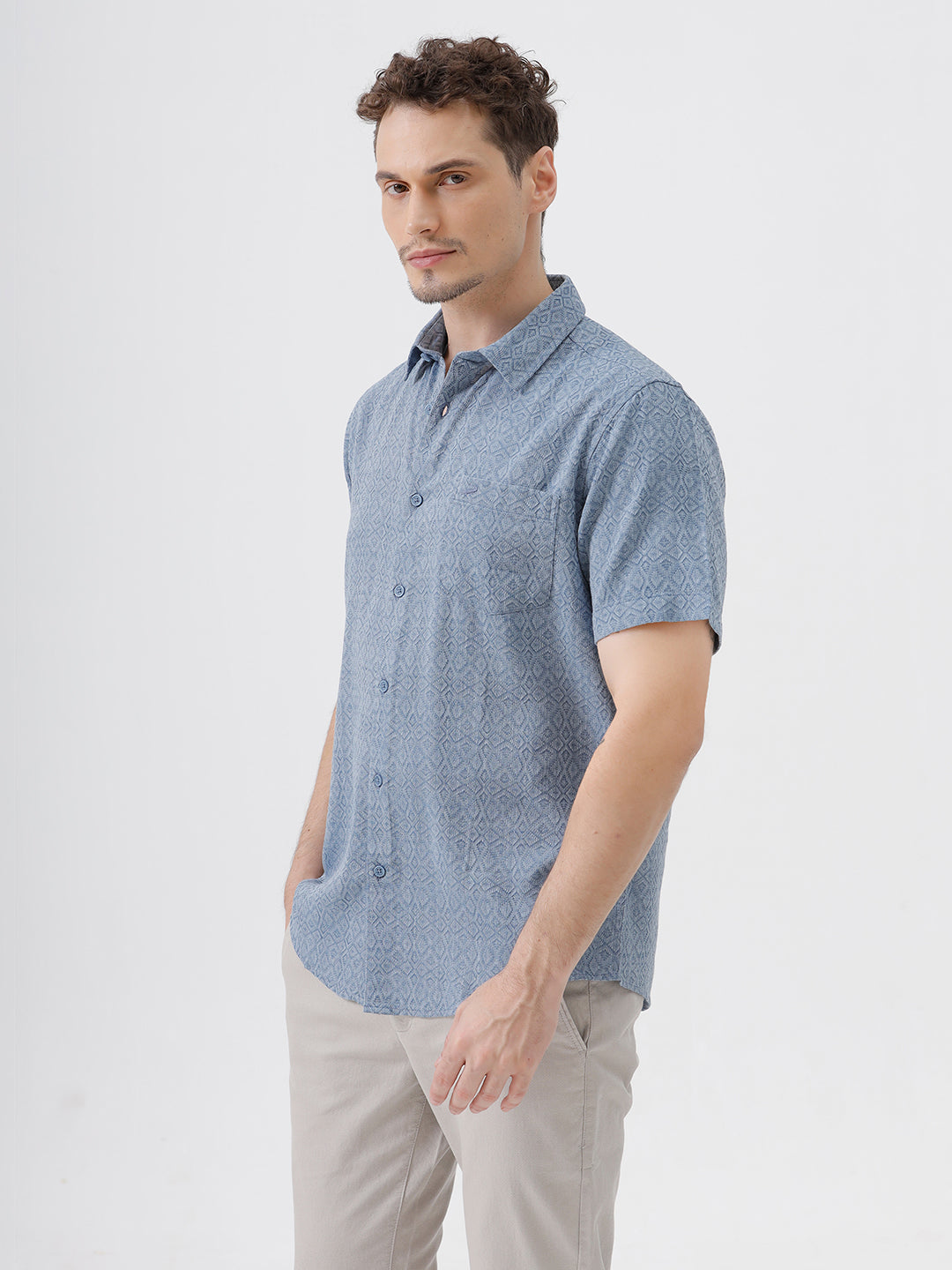 A man wearing a short-sleeve button-up shirt with a fine blue and white geometric pattern, paired with light beige trousers, standing against a plain light gray background.