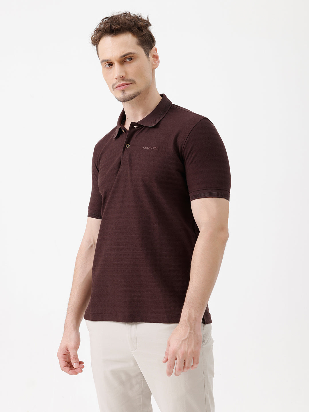 A man wearing a dark brown short-sleeve polo shirt with a subtle horizontal stripe pattern and light beige trousers, standing against a plain white studio background.