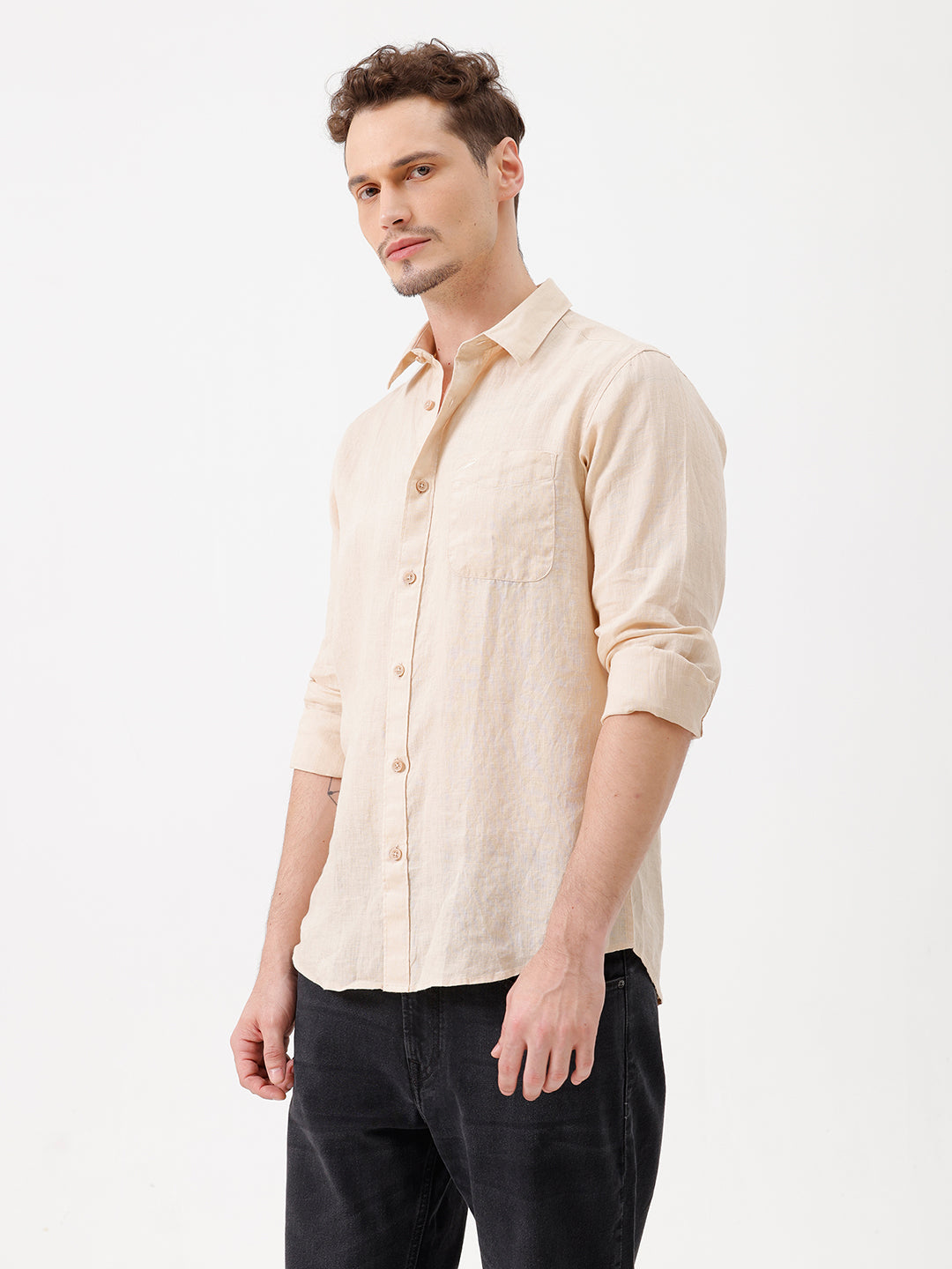 A man wearing a light beige linen button-up shirt with rolled-up sleeves and dark jeans, standing against a plain white studio background.