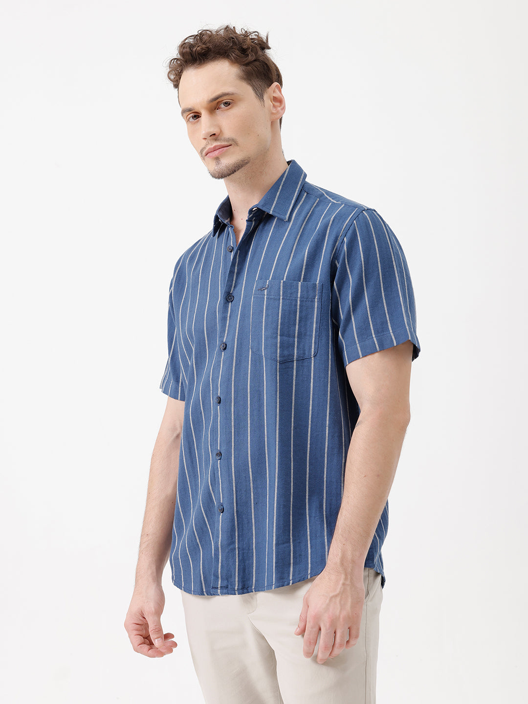 A man wearing a blue short-sleeve button-up shirt with thin white vertical stripes and beige trousers, standing against a plain white studio background.