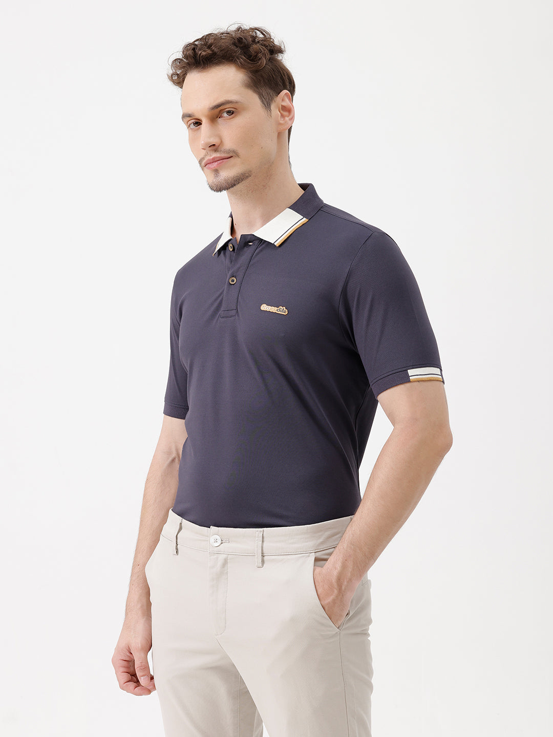 A man wearing a navy blue short-sleeve polo shirt with a contrasting cream and gold striped collar, paired with beige trousers, against a plain white studio background.