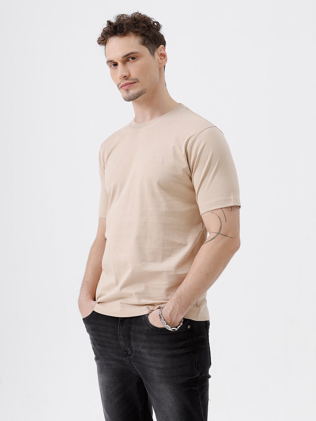 A man wearing a beige short-sleeve crewneck t-shirt with a subtle tonal logo on the left chest, paired with black jeans, standing against a light gray studio background.