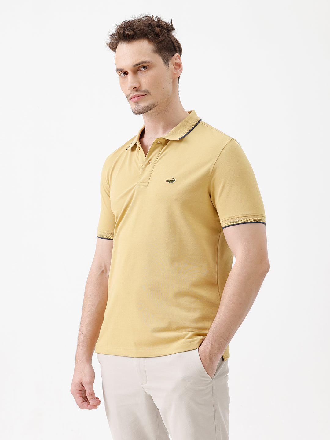 A man wearing a light yellow Lacoste polo shirt with dark piping on the collar and sleeve cuffs, paired with beige trousers, against a plain white studio background.