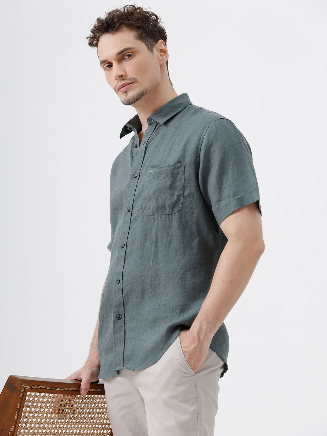 A man wearing a short-sleeve button-up shirt in a muted slate green color, paired with light beige trousers, standing against a plain light gray background.
