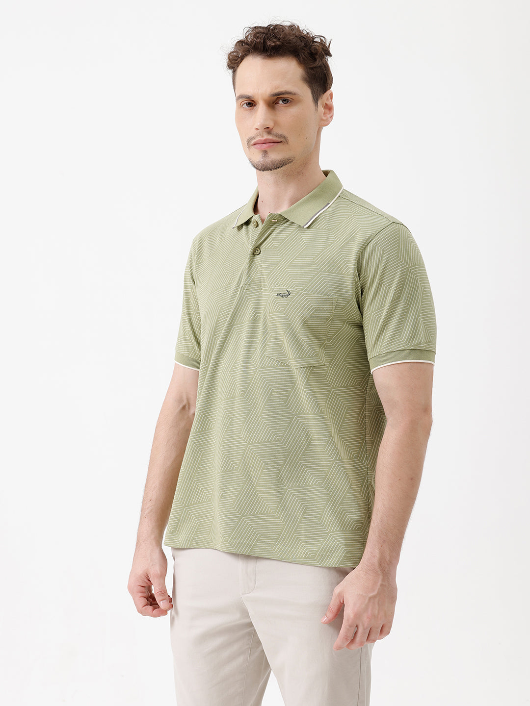 A man wearing a light sage green short-sleeve polo shirt with a subtle geometric pattern, paired with beige trousers, standing against a plain white studio background.