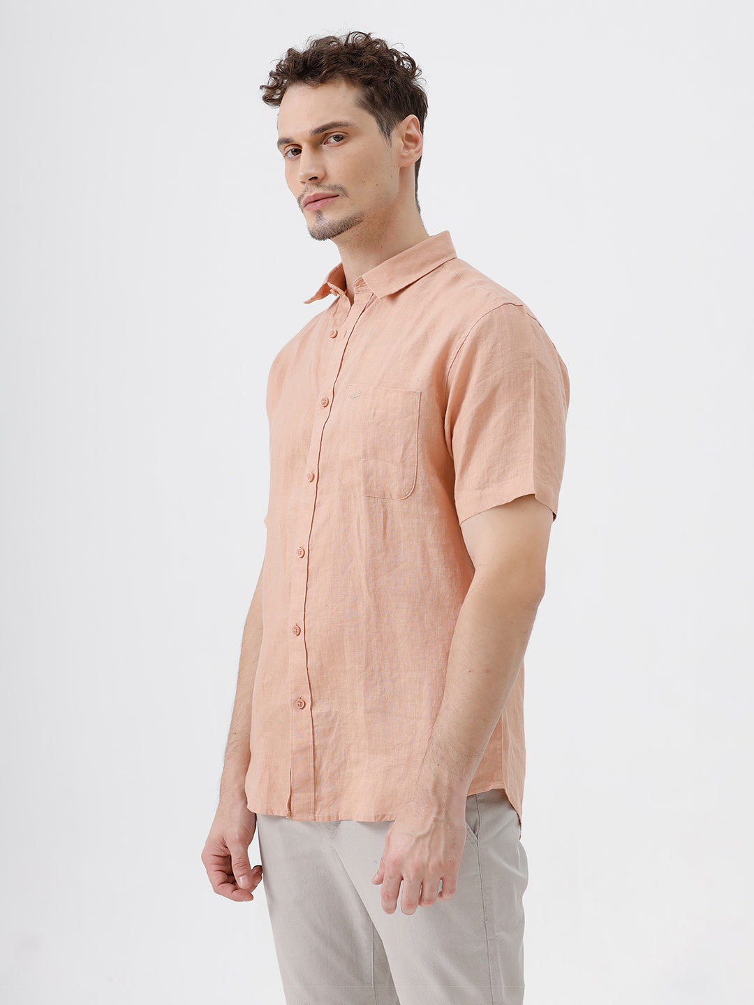 A man wearing a short-sleeve linen button-up shirt in a light peach-pink color, paired with light gray trousers, standing against a plain light gray background.