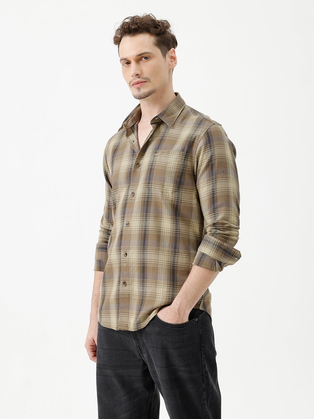 A man wearing a brown and beige plaid long-sleeve button-down shirt with dark sunglasses and black jeans, standing against a plain white studio background.