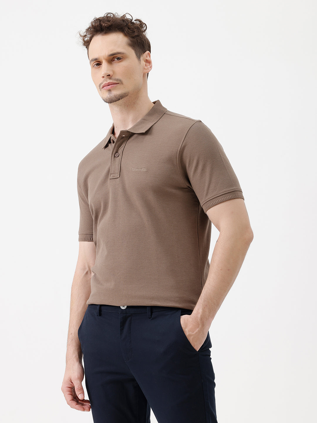 A man wearing a brown short-sleeve polo shirt with dark navy pants, standing against a plain light gray studio background.