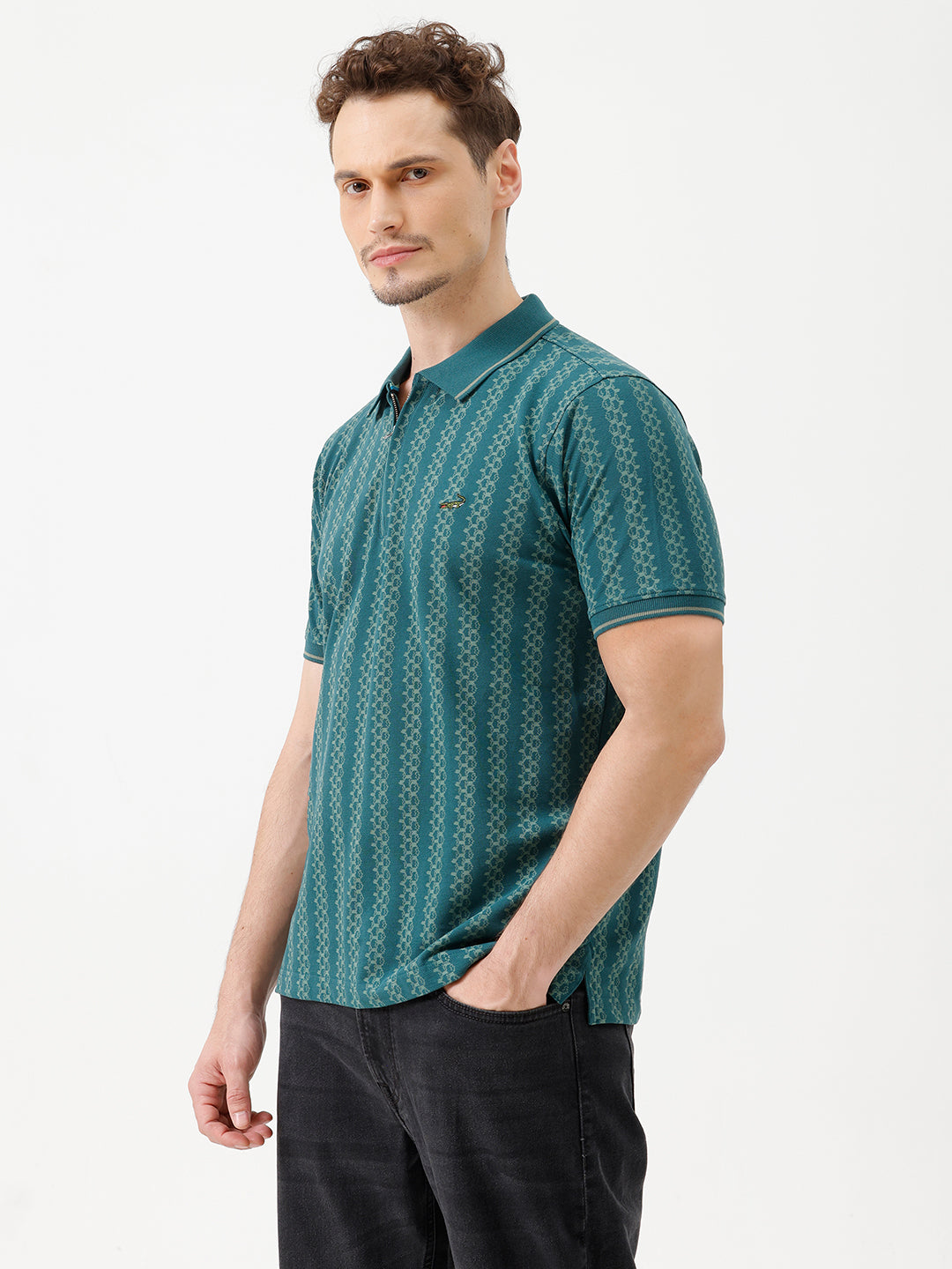A man wearing a teal green short-sleeve polo shirt with a white geometric pattern, paired with black jeans, standing against a plain white studio background.