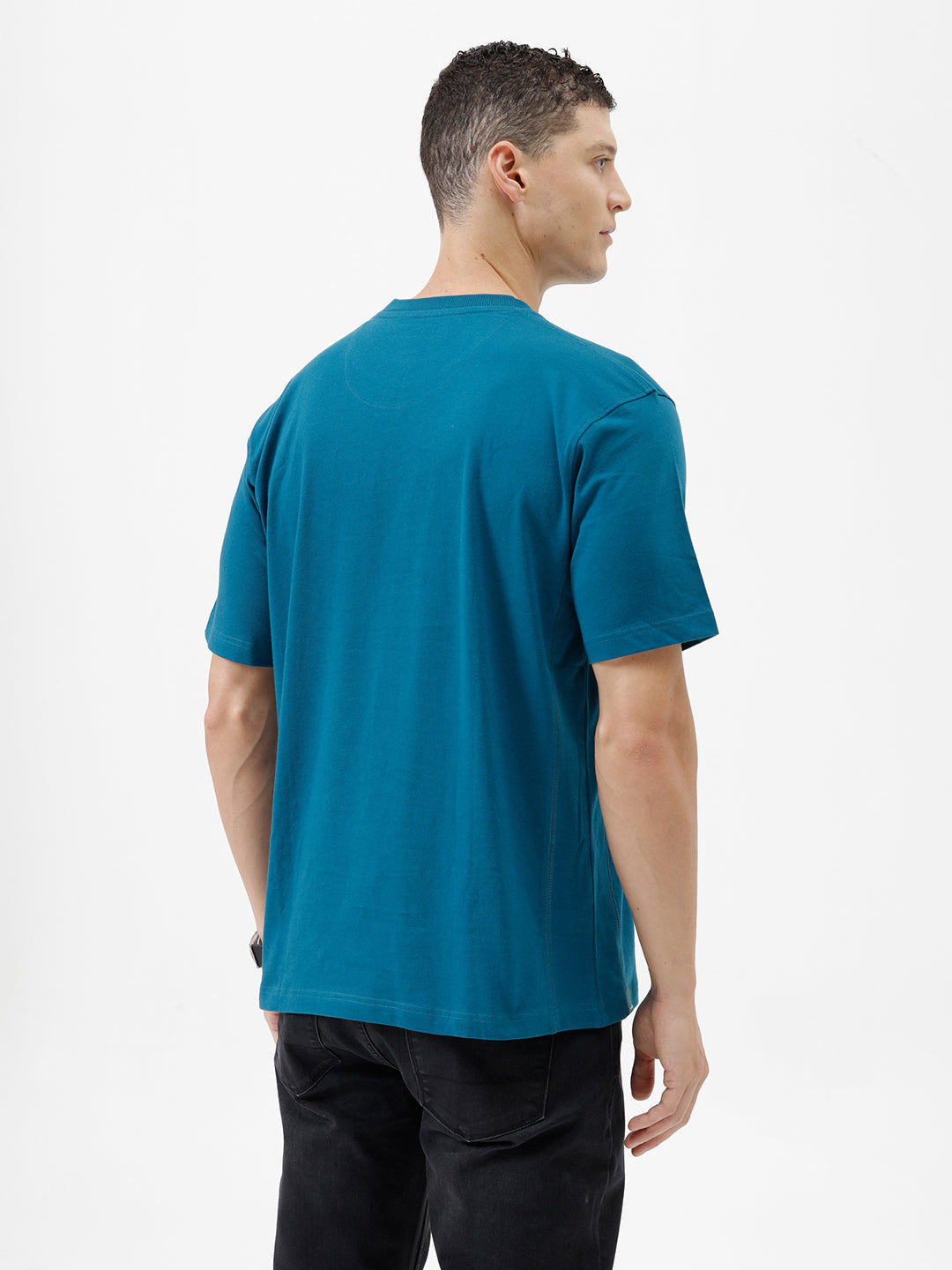 A man wearing a teal blue short-sleeve t-shirt with a subtle tonal 'GREATNESS' text graphic across the chest, paired with black pants, photographed against a clean white studio background.
