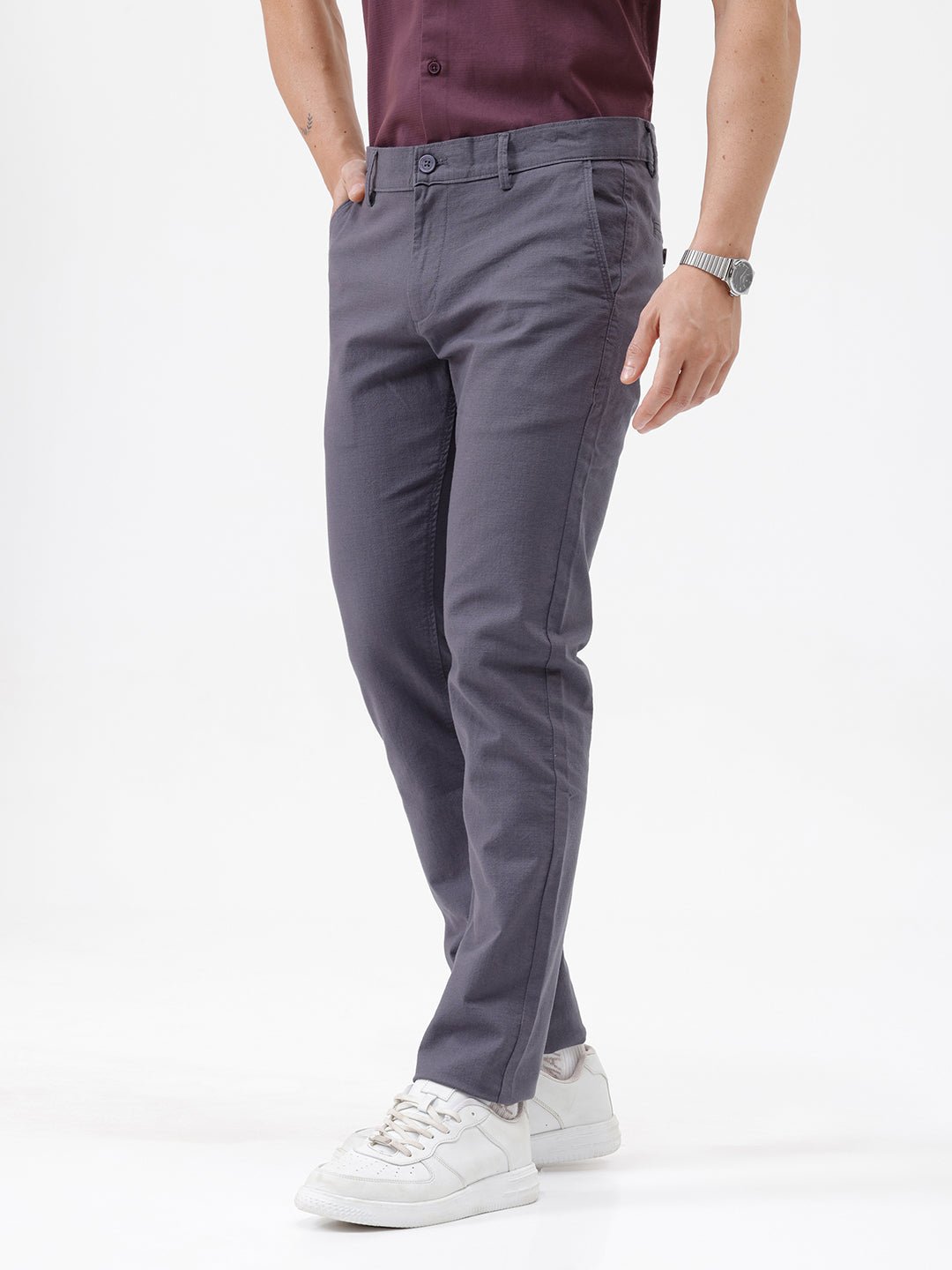 A person wearing slim-fit charcoal gray chino pants, paired with a dark purple shirt and white sneakers, standing against a plain white studio background.