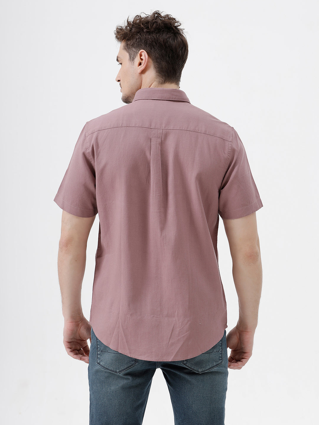 A man wearing a short-sleeve button-up shirt in dusty rose pink with dark blue jeans, standing against a plain white studio background.