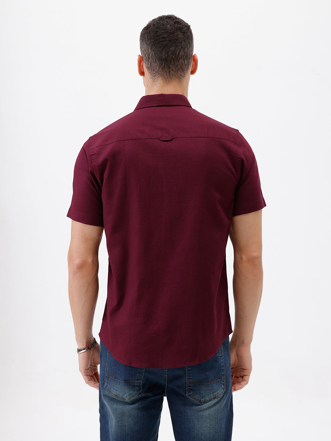 A man wearing a short-sleeve button-up shirt in maroon with a chest pocket, paired with blue jeans, standing against a plain white studio background.