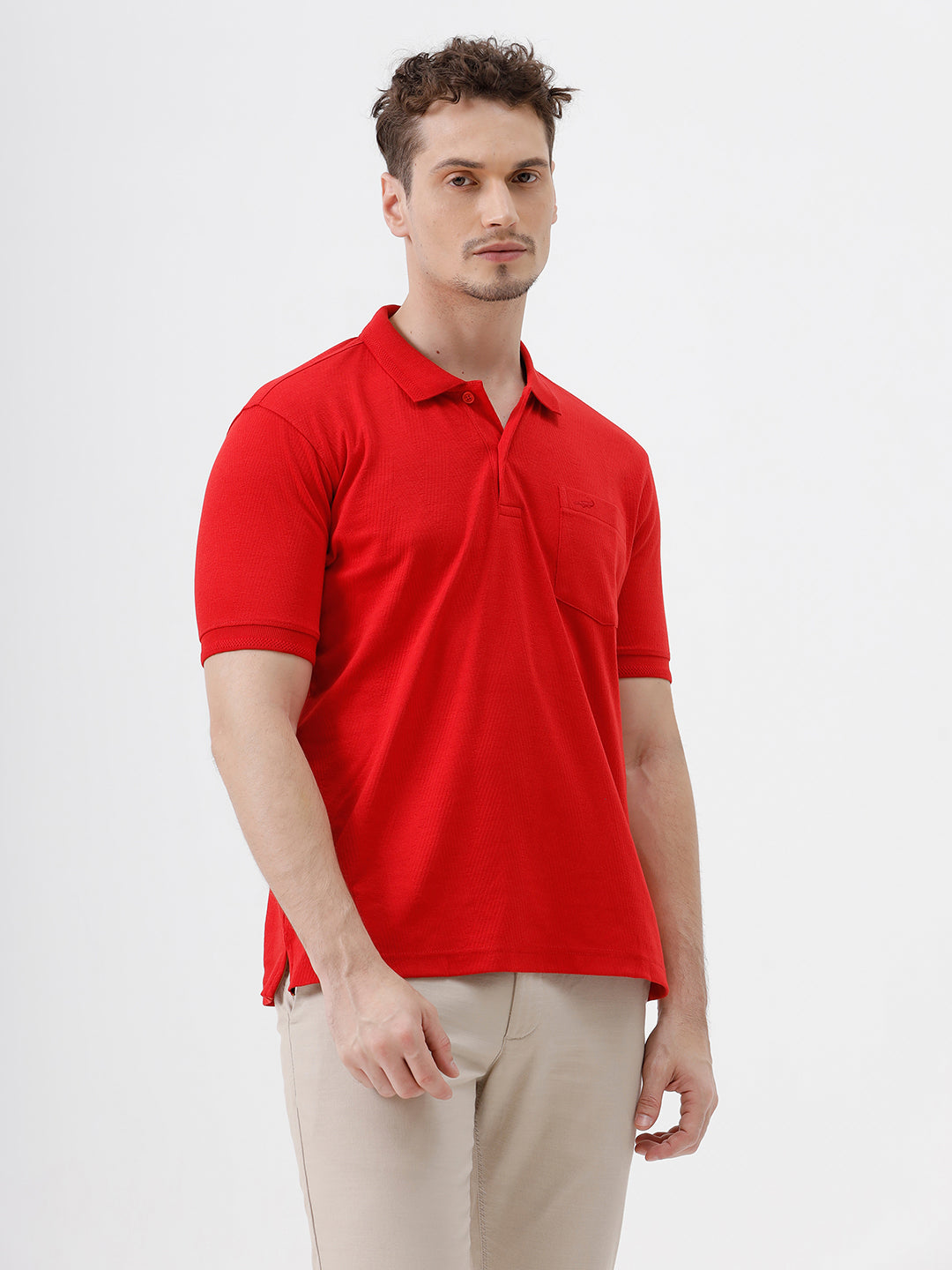 A man wearing a bright red short-sleeve polo shirt with a chest pocket, paired with beige trousers, standing against a plain white studio background.